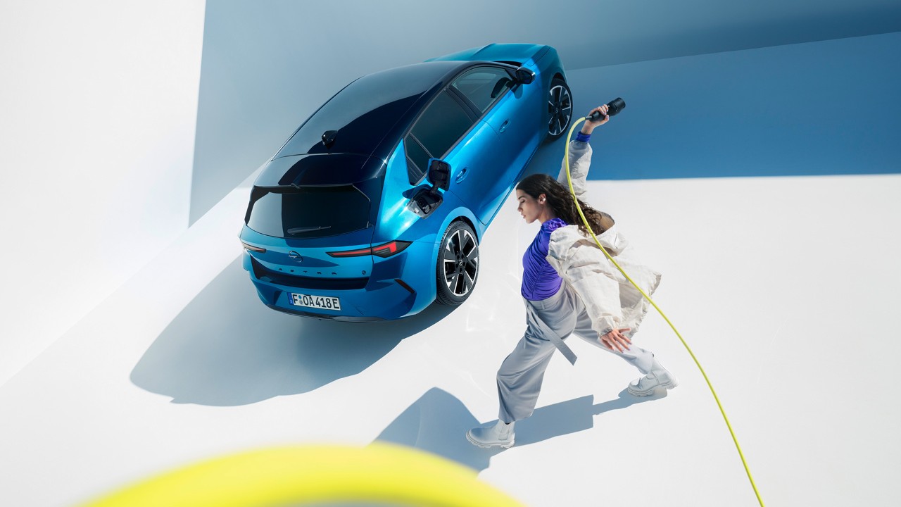 En blå Opel Astra Electric sedd ovanifrån med svart tak, bredvid går en kvinna som snurrar på en gul laddkabel