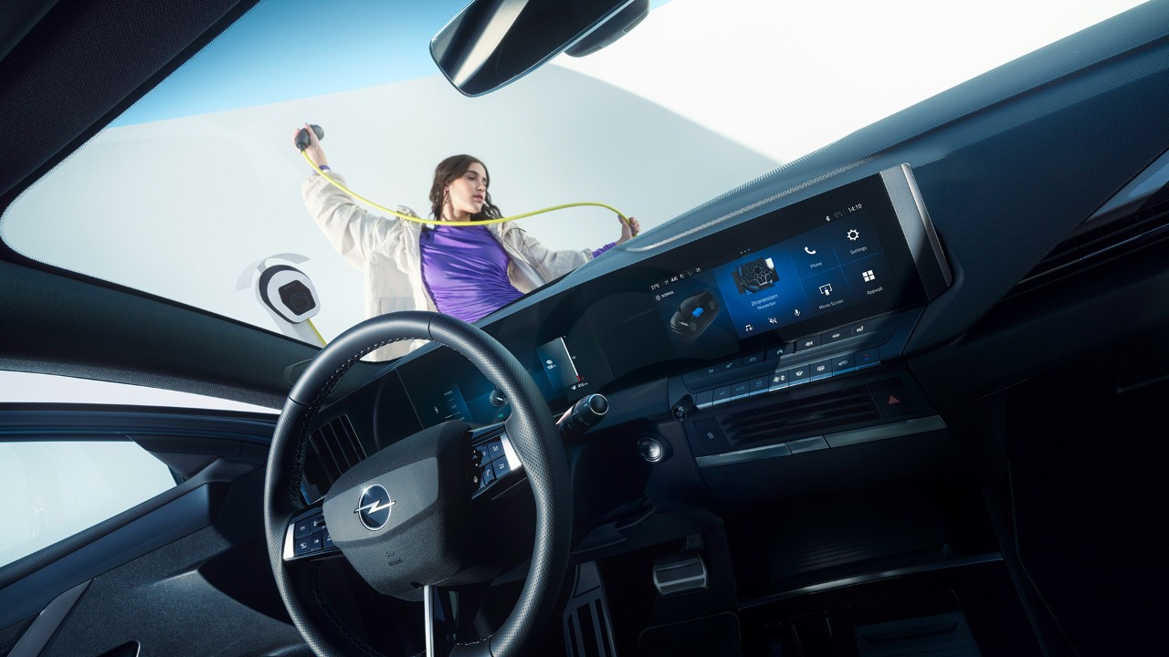 Interiörvy av en Opel Astra Electric från passagerarsätet. Genom framrutan ser man en kvinna som håller en laddningskabel utanför