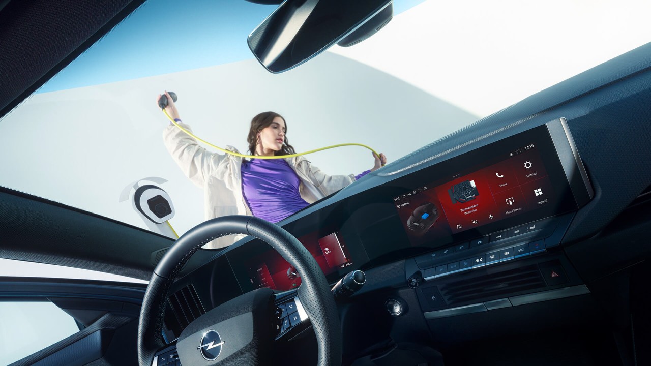 Interiören i Opel Astra Sports Tourer Electric, utanför håller en kvinna med lila tröja en laddkabel