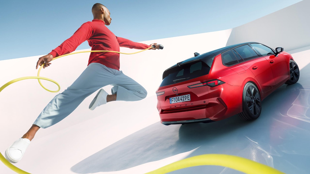 Opel Astra Sports Tourer Electric snett bakifrån, bakom bilen hoppar en man med en laddkabel mot bilen